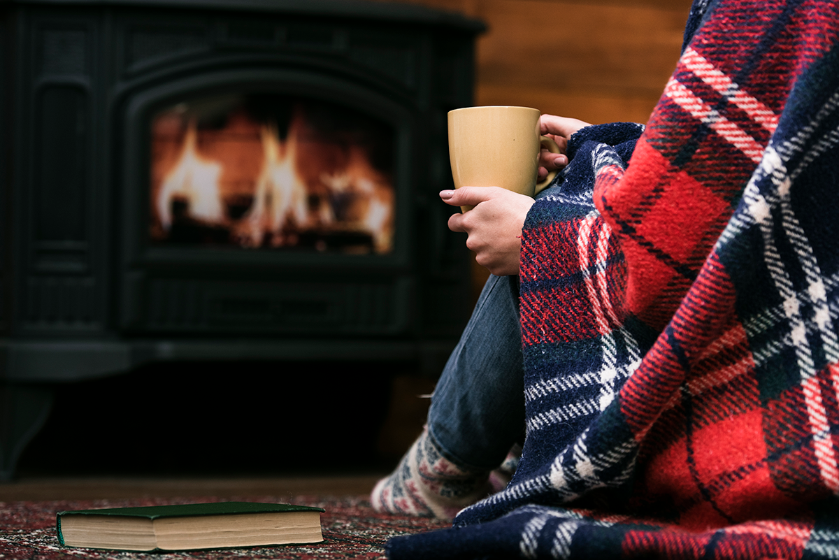 woman with blanket and coffee mug next to fireplace.