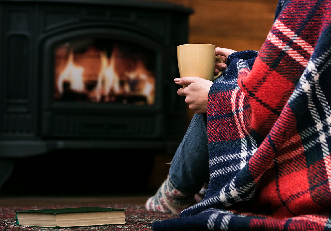 woman with blanket and coffee mug next to fireplace.